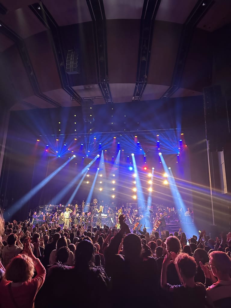 Crowd of concert-goers raising hands before a brightly lit stage with blue and warm spotlights during an orchestra performance.
