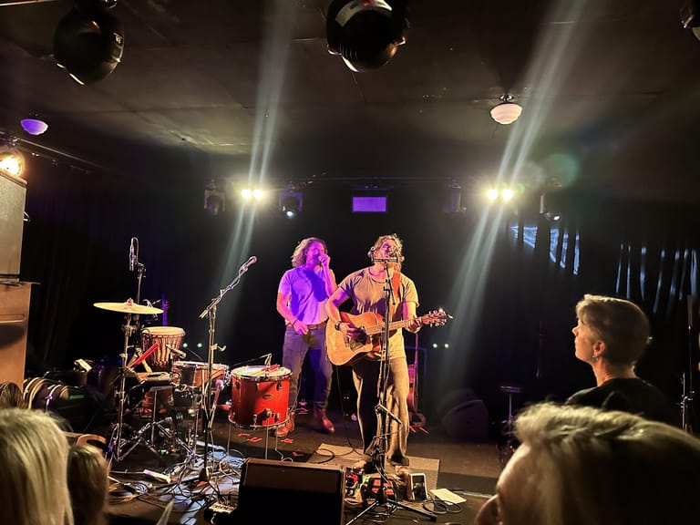 Two musicians perform on stage under bright spotlights, one in purple light playing guitar, the other in warm light with acoustic guitar, with drummer and audience visible.
