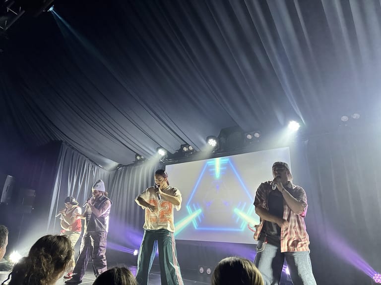 Four performers on stage under bright spotlights with geometric backdrop projection during live concert performance.