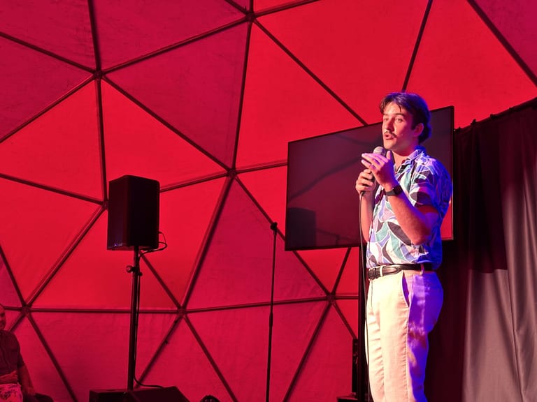 Performer on stage holding microphone against geometric red backdrop lit with purple spotlights.