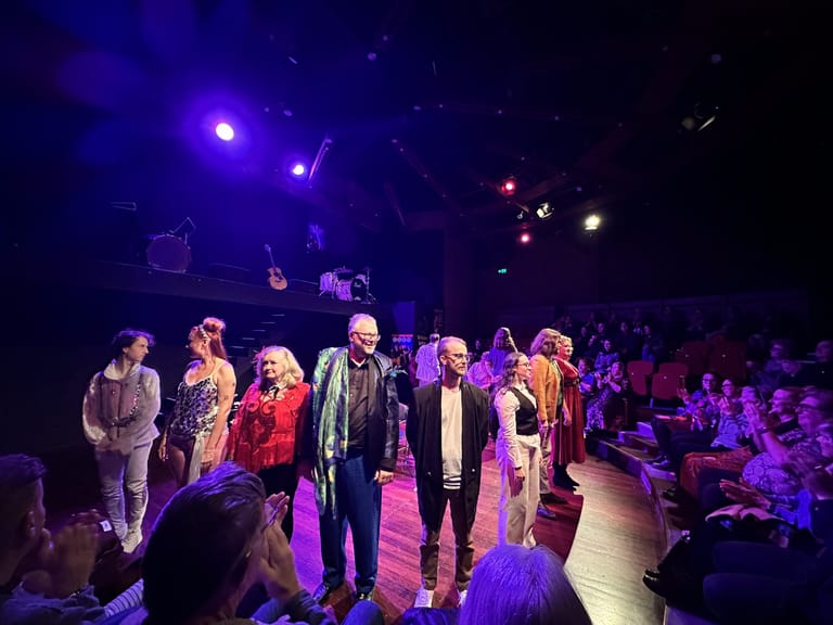 Cast members stand on stage in coloured lighting during a theatrical performance of The Taming Of The Shrew.