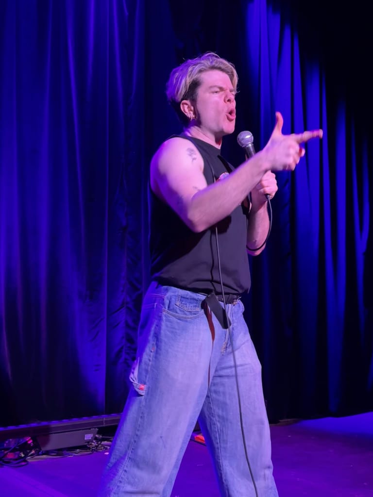Performer on stage holding a microphone with arm extended, wearing a dark sleeveless top and jeans, lit by purple and blue stage lighting.