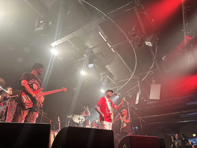 Guitarist in red shirt performs centre stage under red and white spotlights with band members alongside.