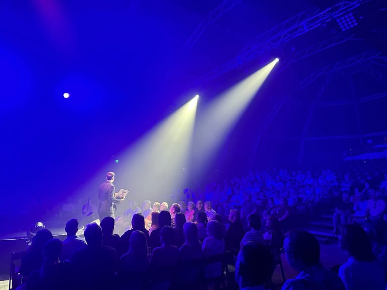 Performers on stage bathed in blue and white spotlights with an engaged crowd in the foreground.