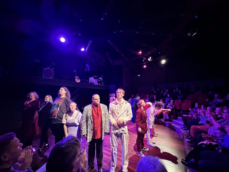 Cast members in period costume stand centre stage under purple and red stage lighting during a theatrical production.