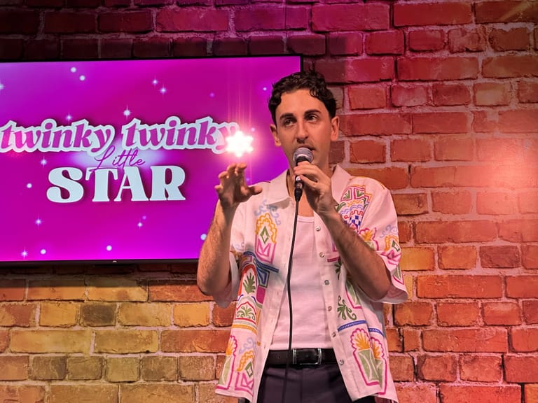 Comedian in a patterned pink jacket speaks into a microphone on stage beneath a glowing neon "Twinky Twinky Little Star" sign.