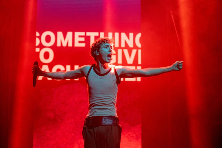 Performer with arms outstretched stands centre stage against a vibrant red backdrop with white text.