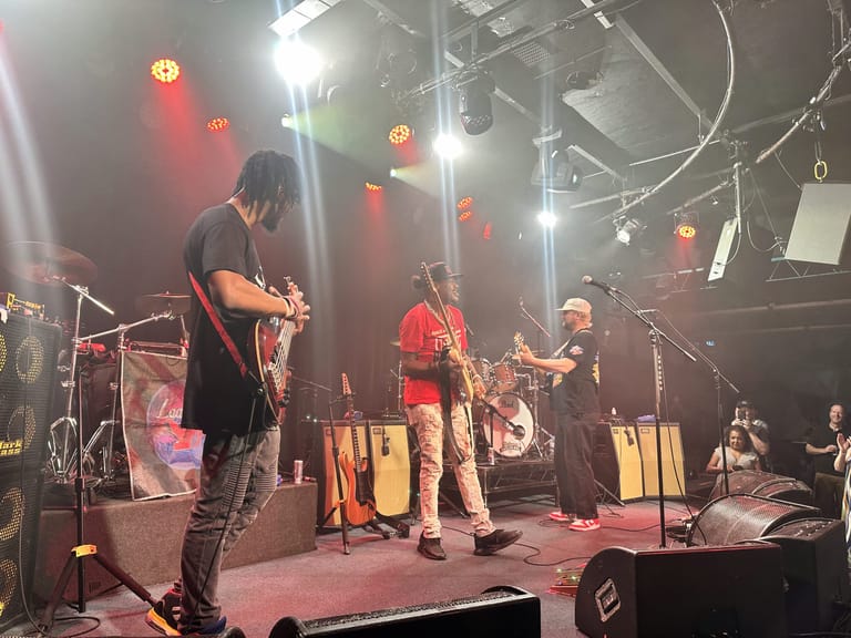 Guitarist in dark shirt performs centre stage with band members under red and white stage lighting.