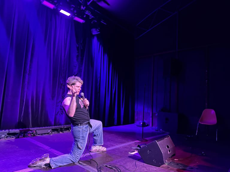 Performer kneeling on stage in purple spotlight with blue curtains and equipment visible behind.