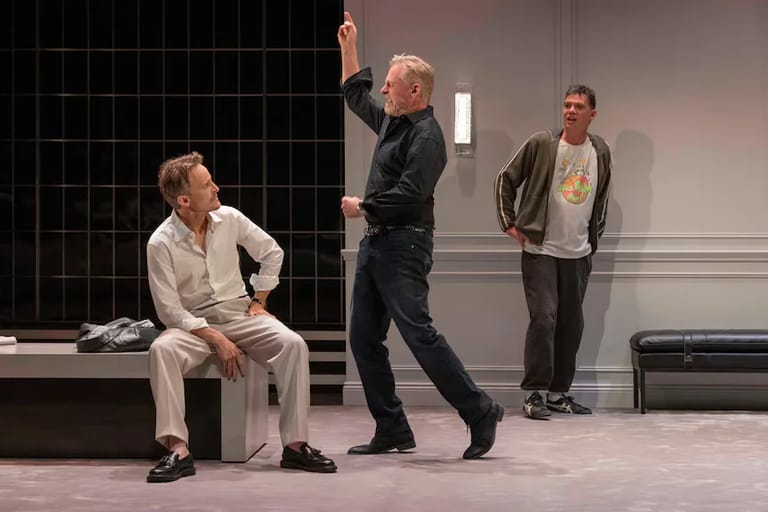 Three male actors on stage during a scene from the play Art, with one seated and two standing in conversation.
