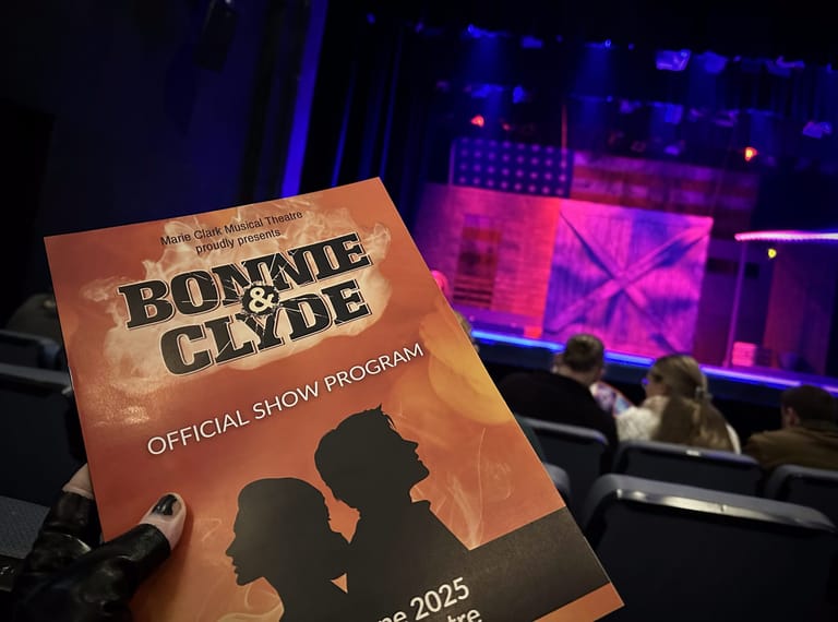 Bonnie & Clyde show programme held in foreground with colourful stage lighting and performers visible in background theatre.