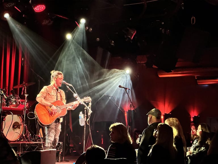 Musician with acoustic guitar performing on stage under dramatic red and white lighting with audience silhouettes in foreground.