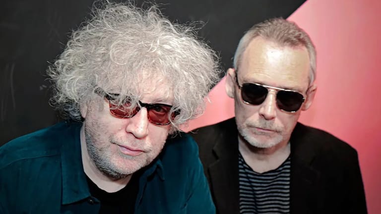 Two men wearing sunglasses pose against a black and pink background during an interview.