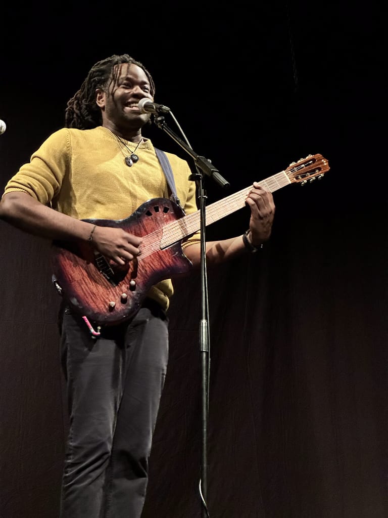 Performer in yellow shirt playing acoustic guitar at microphone against dark stage background.