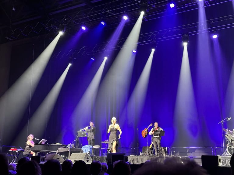 Male singer in yellow-green shirt centre stage, flanked by band members under dramatic blue and white spotlights.