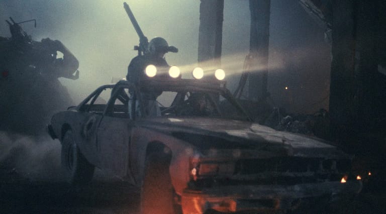 Wrecked car with glowing lights and robotic figure in smoky, industrial post-apocalyptic setting.