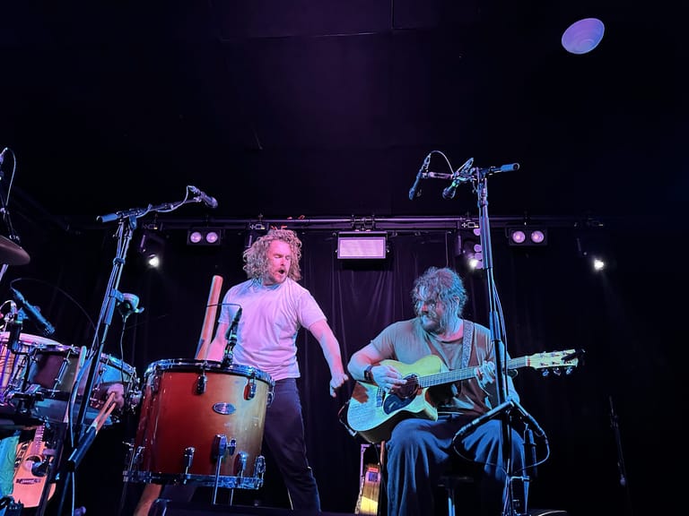 Two musicians perform on stage under purple lighting, one singing while the other plays guitar, with drum kit visible to the left.