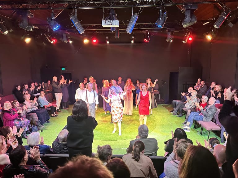 Performers and audience members on a brightly lit green stage floor surrounded by seated spectators in an intimate theatre setting.