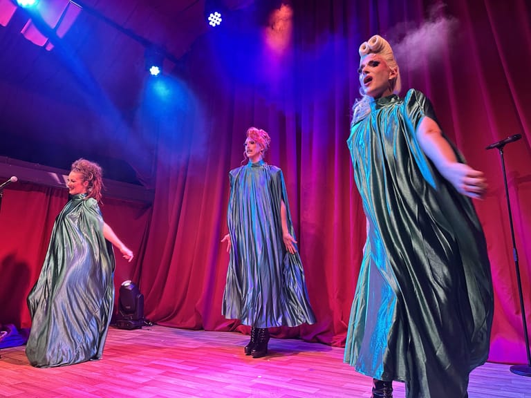 Three performers in shimmering pleated gowns stand on a pink-lit stage under blue and purple spotlights during Mass Hysteria.