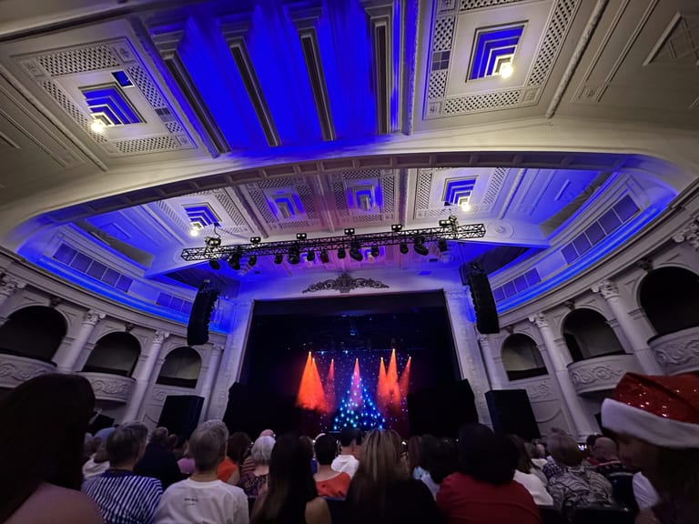 Theatre ceiling and stage lit in blue and red, with audience watching a Christmas-themed performance.