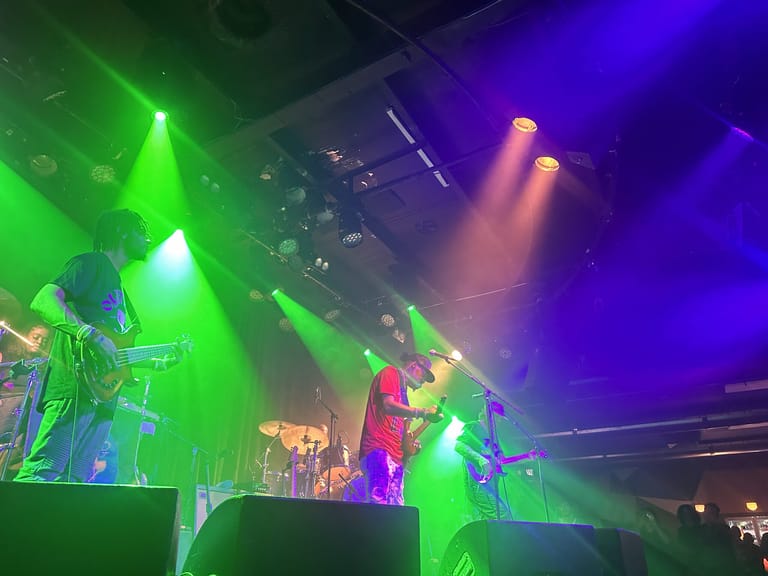 Guitarist performing on stage bathed in vibrant green and purple stage lighting during a live concert.