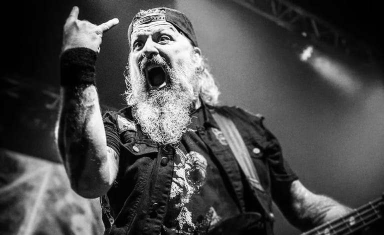 Bearded performer with long hair singing passionately into microphone on darkened stage, fist raised.