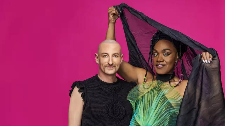 Two performers pose together against a bright magenta background, holding a black fabric between them.