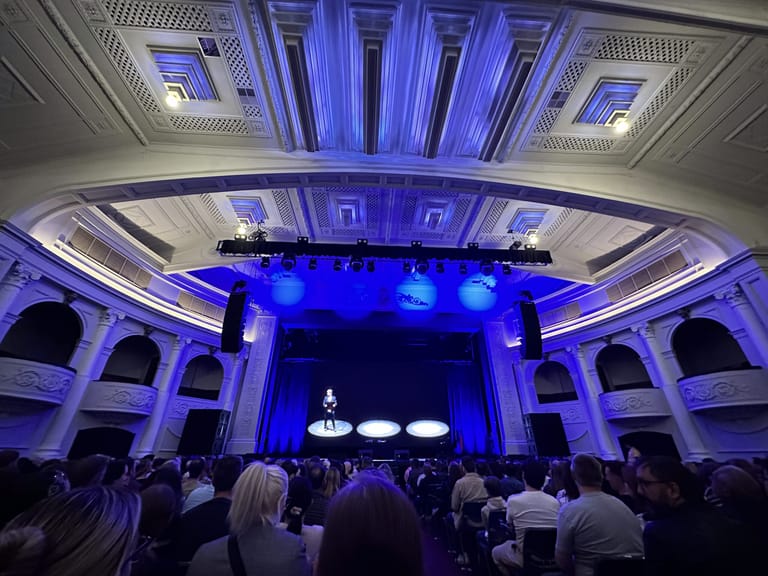Performer on stage in a blue-lit theatre with ornate ceiling, audience seated in foreground.