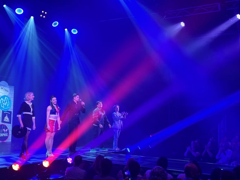 Five performers stand centre stage bathed in blue and magenta spotlights during a theatrical production.