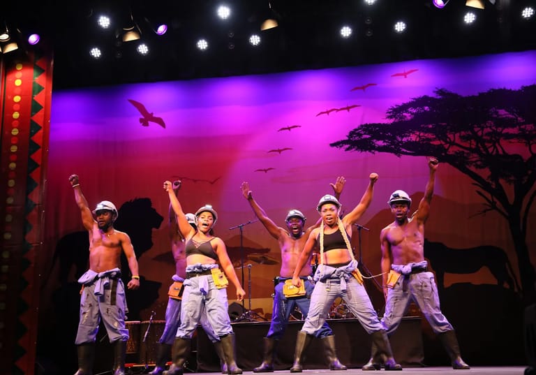 Four shirtless male dancers in white cargo pants perform with raised arms on a stage bathed in purple and pink lighting.