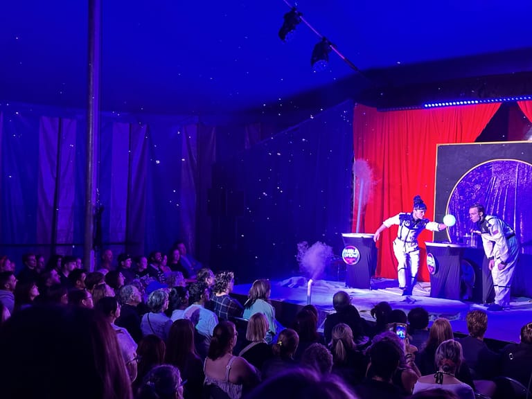 Performer in striped costume creates large bubbles on stage as audience watches under blue and red stage lighting.