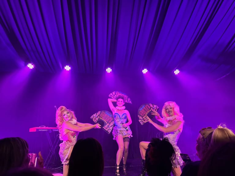 Four performers in sequined costumes dance under purple stage lights during a cabaret production.