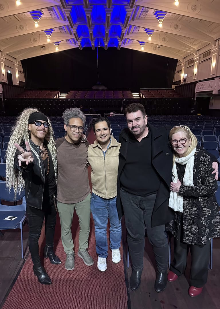Five band members stand together on an empty theatre stage with blue uplighting and architectural details visible above.
