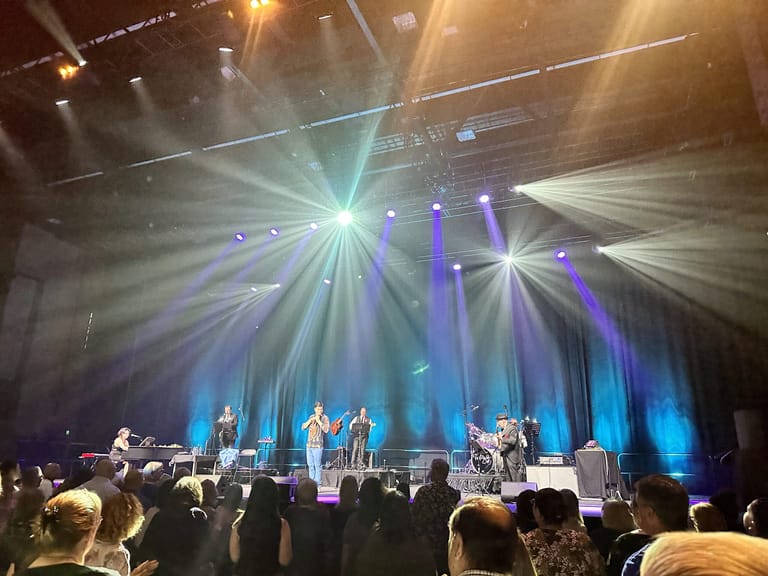 Performer in orange shirt centre stage surrounded by band, dramatic blue and white stage lighting illuminating audience silhouettes.