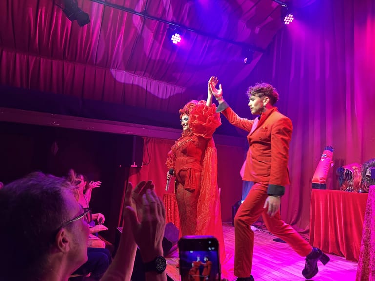 Two performers in red outfits engage with audience members on stage under vibrant purple lighting.
