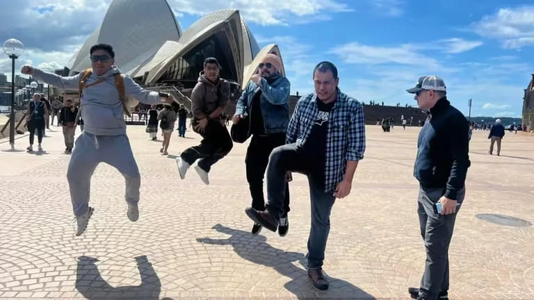Five musicians jumping in unison on a sunny plaza with the Sydney Opera House in the background.