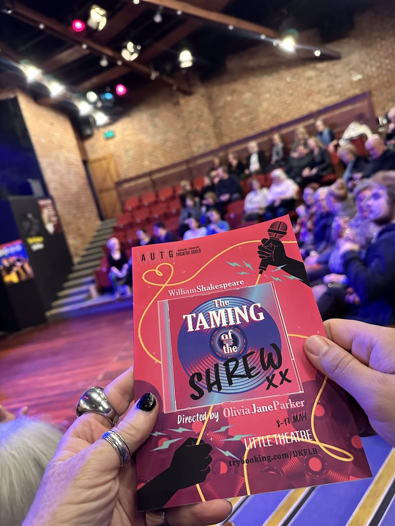 Hand holding a red programme for The Taming of the Shrew at an intimate theatre venue with audience seated in background.