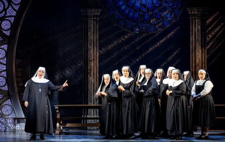 A nun conducts a choir of nuns in period costume on a theatrical stage with ornate architectural backdrop.