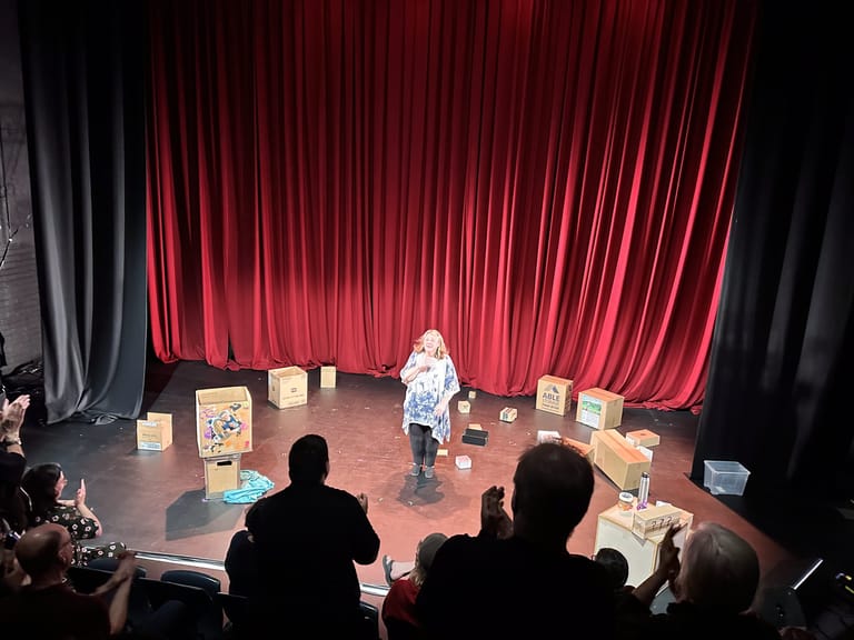 Performer stands centre stage surrounded by cardboard boxes, framed by dramatic red curtains and coloured stage lighting.