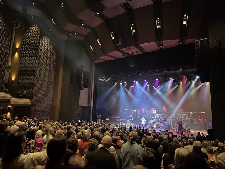 Packed theatre audience facing an illuminated stage with band and performers under coloured spotlights.