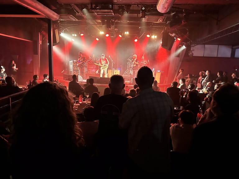 Band performing on stage bathed in red lighting, with silhouetted audience members in the foreground.
