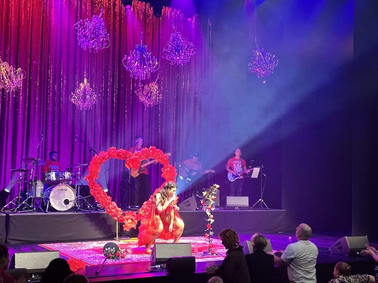 Performer on stage within a large illuminated heart prop, surrounded by purple lighting and hanging crystal decorations, with band members and audience visible.