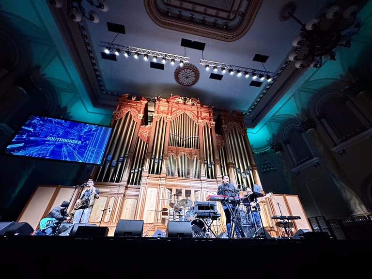 Large pipe organ illuminated in warm light with blue stage lighting and drum kit in foreground during concert performance.