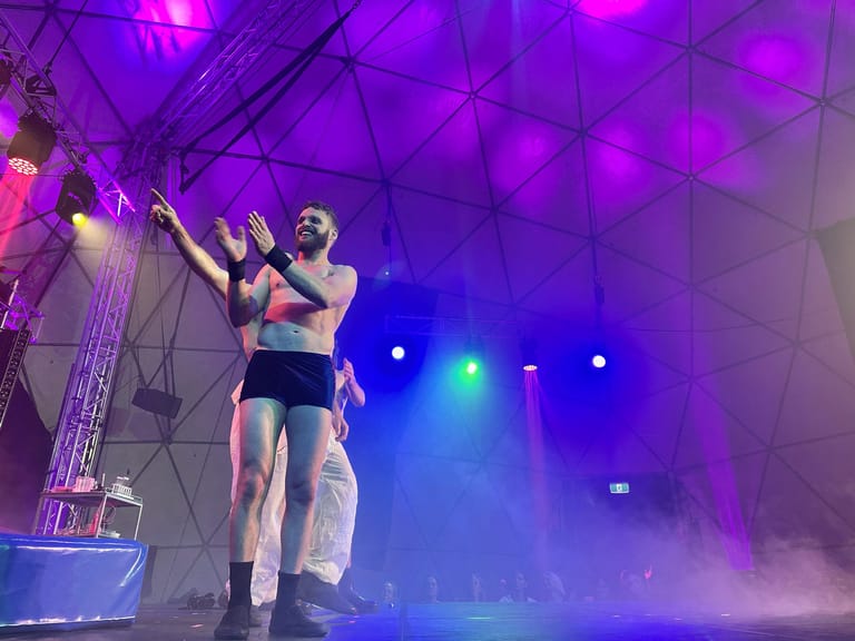 Performer in black crop top and shorts strikes a pose on stage amid purple and blue lighting and stage fog.