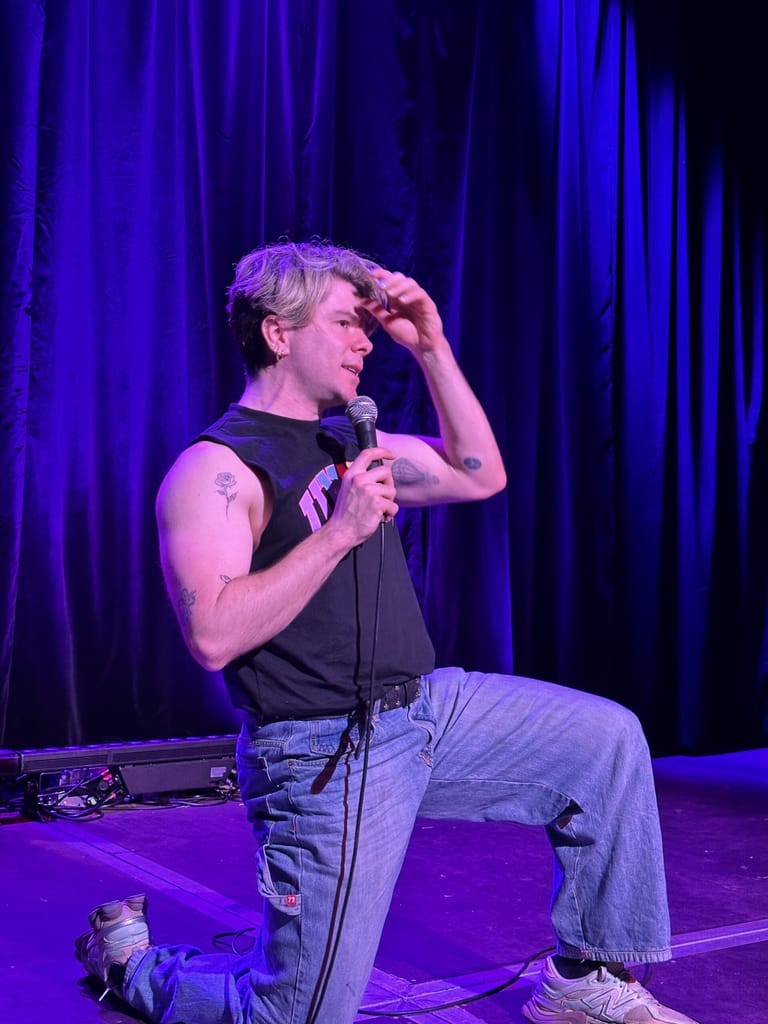 Performer on stage holding a microphone, lit by purple spotlights against a dark backdrop.