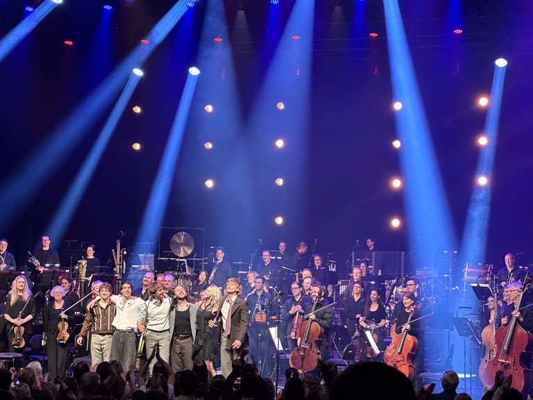 Lime Cordiale and orchestra members stand together on stage beneath dramatic blue concert lighting.