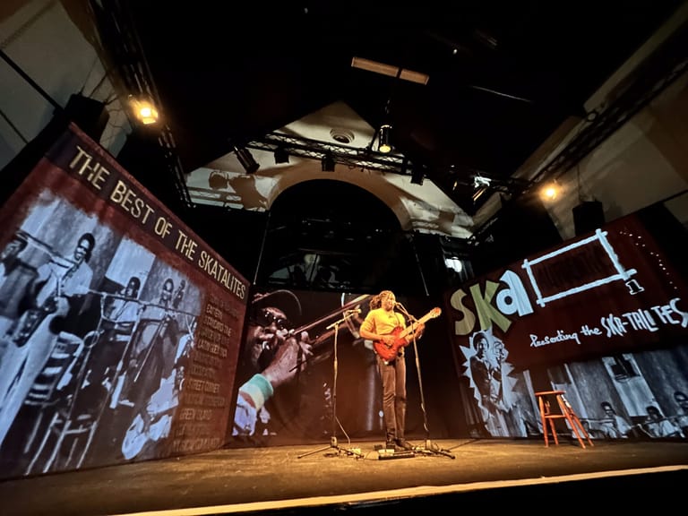 Performer with dreadlocks and yellow shirt stands on stage under warm spotlights, surrounded by projected images and vintage photographs on brick walls.