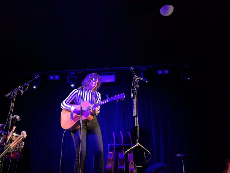 Musician with acoustic guitar centre stage under purple spotlights during live performance.