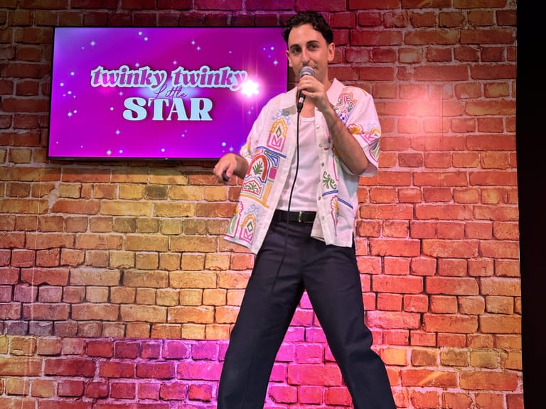 Comedian performs on stage before a neon purple "twinky twinky STAR" sign on a brick wall.
