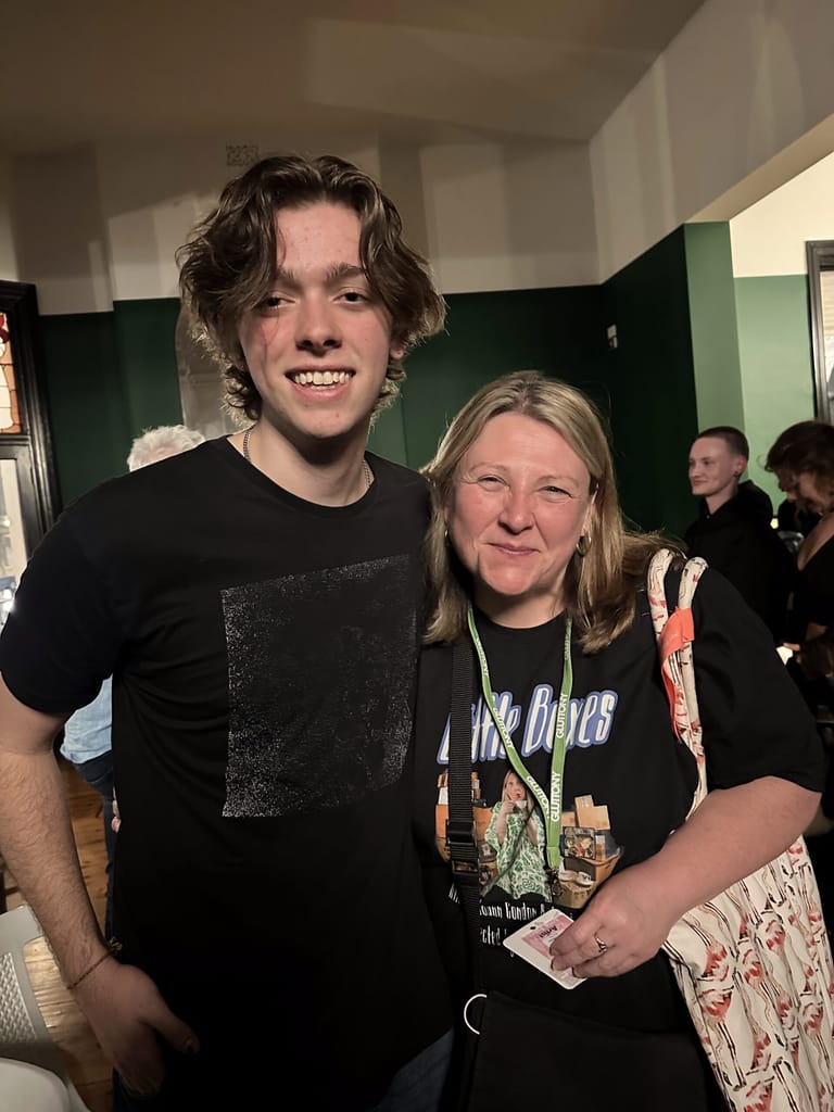 Two people smile at the camera in a green-walled venue, with other attendees visible in the background.